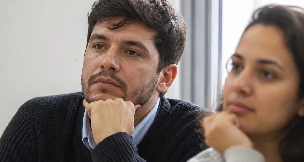 Participante com o braço no alto pedindo a vez, diante de folhas rabiscadas espalhadas pela mesa